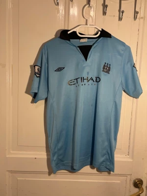 Manchester City matchtröja ljusblå (2012-13) - Säljes snabbt, är öppen för seriösa bud.        Ljusblå Manchester City matchtröja från Umbro med svart krage och broderat klubbmärke på bröstet. Tröjan har Etihad Airways som sponsortryck och Premier League-märke på ärmen. Perfekt för fotbollsfans som vill visa sitt stöd på läktaren eller planen.