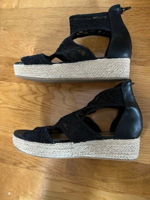 Svarta kilklackade sandaler med spets - Snygga svarta sandaler med kilklack och flätad sula i rep. Ovandelen är i spets och konstläder med coola utskärningar och dragkedja bak. Perfekta för att lyfta din sommarstil med en trendig touch.