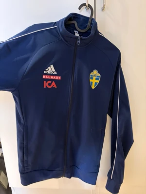Blå Adidas Sverige träningsjacka - Mörkblå träningsjacka från Adidas med Sveriges landslagsemblem på bröstet. Jackan har dragkedja, ståkrage och vita detaljer längs ärmarna. Sponsorerna Bauhaus och ICA är tryckta på framsidan. Perfekt för träning eller till vardags. 