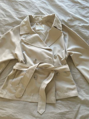 Beige trenchcoat från Stradivarius - Stilren beige trenchcoat från Stradivarius med klassisk dubbelknäppning och knytskärp i midjan. Jackan har bred krage och två sidofickor. Tillverkad i ett mjukt tyg som ger en snygg siluett och passar perfekt till lager på lager.