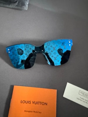Blå Louis Vuitton solglasögon - Snygga solglasögon från Louis Vuitton med blå spegelglas och ikoniskt LV-monogram över hela linsen. Bågarna är mattsvarta med LV-logga på sidan. Glasögonen har en modern, kantig form och känns riktigt exklusiva.