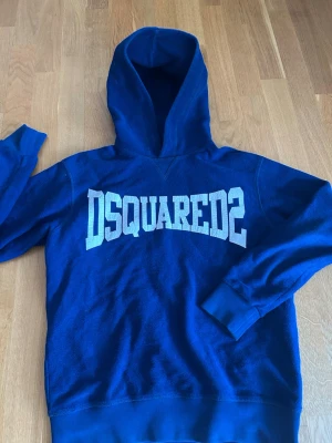 Blå hoodie från Dsquared2 - Snygg blå hoodie från Dsquared2 med stor vit logga över bröstet. Klassisk passform med huva och ribbade muddar vid ärmslut och nederkant. Perfekt för dig som gillar streetwear och vill ha något bekvämt och trendigt. Materialet känns mjukt och skönt mot huden. Skriv vid fundering eller prisförslag😃