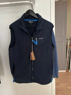 Mörkblå fleeceväst från Patagonia - Mörkblå fleeceväst från Patagonia med dragkedja framtill, två sidofickor och en bröstficka med blå detaljer. Västen har hög krage och klassisk passform, perfekt för lager-på-lager. Ikonisk Patagonia-logga på bröstet. Passar även M 