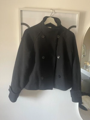 Svart kort rock med hög krage - Stilren svart rock med hög krage och dubbelknäppning framtill. Jackan har långa ärmar med dekorativa spännen vid ärmsluten och en rak, något kortare passform. Perfekt för dig som gillar en klassisk men modern look.