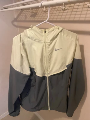 Nike Miller grön,grå Storlek S - Snygg vindjacka från Nike i tvåfärgad design med grön upptill och mörkgrå nedtill. Jackan har hel dragkedja, huva och Nike-logga på bröstet. Perfekt för blåsiga dagar och sportiga outfits.