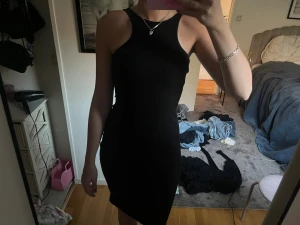 Svart tight ärmlös klänning - Säljer en svart, tight och ärmlös klänning med hög halsringning och öppen rygg. Klänningen är kort och har en stilren, minimalistisk design som passar perfekt till olika tillfällen. Materialet känns mjukt och följsamt mot kroppen.