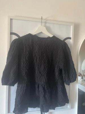 Svart strukturerad blus med knytband - Svart blus från Monki med strukturerat tyg och knytband både vid halsen och midjan. Blusen har en lös passform och trekvartsärmar med lätt puff. Perfekt för en trendig och avslappnad look.