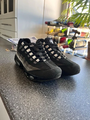Nike Air Max 95 svarta sneakers - Säljer ett par Nike Air Max 95 sneakers i svart med grå detaljer. Skorna har en ikonisk vågformad design, synlig Air-dämpning i sulan och mesh- samt mockadetaljer upptill. Perfekta för dig som gillar streetwear och sportig stil.