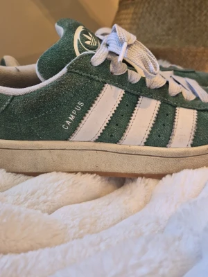 Adidas Campus gröna sneakers - Gröna Adidas Campus sneakers i mocka med klassiska vita ränder på sidorna och vit logga på plösen. Skorna har vita skosnören och en beige sula som ger en retrovibe. Perfekta för dig som gillar streetwear och tidlös stil.