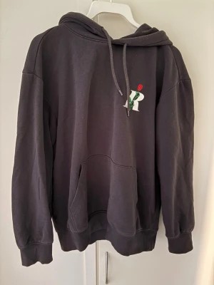 Svart hoodie  - Snygg svart hoodie från hm med broderad logga och röd ros på bröstet. Klassisk känguruficka framtill, ribbade muddar och justerbar dragsko i huvan. Perfekt för en avslappnad och trendig look.