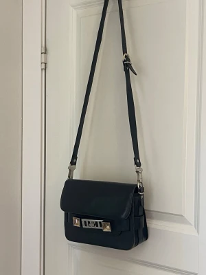 Svart axelväska från Proenza Schouler - Snygg svart axelväska i läder från Proenza Schouler med justerbar axelrem, silverfärgade metalldetaljer och klassiskt spänne framtill. Väskan har en stilren, rektangulär form och praktiska fack både fram och bak. Perfekt för dig som gillar stilren design.