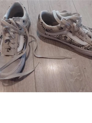Vans Old Skool med döskallemönster - Säljer ett par Vans Old Skool sneakers i vitt och beige med unikt svart döskallemönster på sidorna. Klassisk vit sidestripe och gummisula i brunt. Ovandelen är i canvas och mocka, med vita skosnören och lågt skaft. Perfekta för dig som gillar streetstyle!