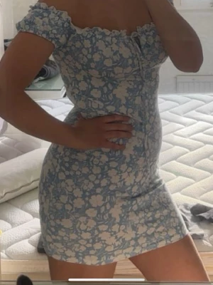 Blå blommig kortklänning med volang - Söt kortklänning i ljusblått med vitt blommönster och volangdetaljer vid axlarna. Klänningen har offshoulder-modell och knytning framtill. Perfekt för varma dagar och har en feminin, figurnära passform.