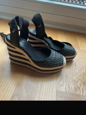 Svarta kilklackade espadrilles med snörning 38 - Snygga svarta espadrilles med kilklack och snörning runt ankeln. Ovandelen är i flätad textil och sulan har ett randigt mönster i svart och naturfärgad jute. Perfekta för en trendig sommarlook.