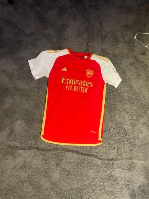 Arsenal matchtröja Adidas röd/vit - Passar som M Säljer en  Arsenal matchtröja från Adidas i rött och vitt med gula detaljer. Tröjan har klubbmärke och sponsortryck på bröstet samt korta ärmar. Perfekt för fotbollsträning eller att bära på matchdag. Materialet är lätt och andas.