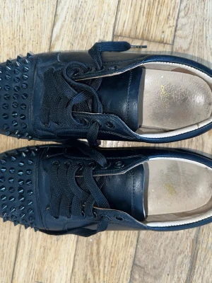 Svarta sneakers med nitar från Louboutin - Säljer ett par svarta sneakers från Christian Louboutin i läder med svarta nitar över tån och svarta skosnören. Skorna har en låg siluett, säljer den biligare då det saknas 1 spik på varje fot