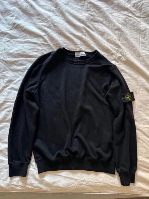 Svart sweatshirt från Stone Island - Svart sweatshirt från Stone Island med klassisk rund halsringning och ribbade muddar. Tröjan har den ikoniska Stone Island-loggan på vänster ärm. Perfekt för en stilren och avslappnad look.