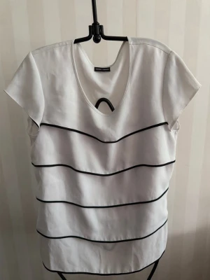 Vit topp med svarta ränder Gerry Weber - Vit topp från Gerry Weber med svarta vågiga ränder och rund halsringning. Kortärmad och i mjukt material som känns skönt mot huden. Perfekt för dig som gillar enkel men snygg design.