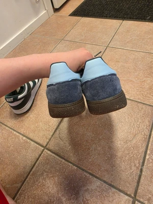 Adidas blå sneakers i mocka - Snygga Adidas sneakers i blå mocka med ljusblå detaljer och klassiska vita ränder på sidan. Skorna har en brun gummisula med mönster för bra grepp. Perfekta för dig som gillar retrostil och vill ha något unikt till din outfit.