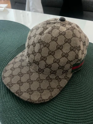 Gucci keps  - Gucci-keps i klassiskt GG canvas med den ikoniska web-detaljen på sidan. En riktigt snygg statement-piece som ger en clean men lyxig känsla till vilken outfit som helst.  Färgerna är lätta att matcha och modellen sitter väldigt fint på. Använd men fortfarande i fint skick, med normalt slitage från användning.  Säljer eftersom den inte kommer till användning längre. Perfekt om du vill få in en tidlös designerdetalj i garderoben utan att betala nypris.  Skriv om du vill ha fler bilder eller mått