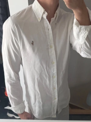 Vit skjorta från Ralph Lauren - Klassisk vit skjorta från Ralph Lauren med button-down krage och broderad logga på bröstet. Skjortan har lång ärm, normal passform och är tillverkad i mjuk bomull. Perfekt till jeans eller chinos för en clean och stilren look. Den är lite missfärgad i kragen och armhålorna, därav priset. Annars bra skick 