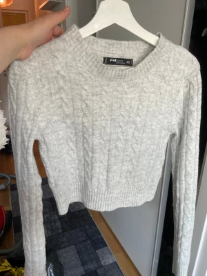 Ljusgrå kabelstickad tröja XS - Ljusgrå kabelstickad tröja från Funky Buddha med croppad passform och rund halsringning. Tröjan har långa ärmar och ribbade muddar vid ärmslut och nederkant. Perfekt för lager-på-lager och en chill look.