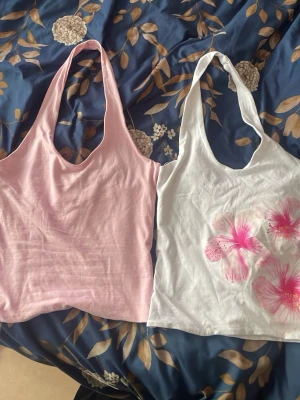 2 linne-toppar i rosa och vitt - Två linne-toppar, en i ljusrosa och en i vitt med rosa blommor. Båda har djup urringning och är ärmlösa med smala axelband. Perfekta för varma dagar och enkla att matcha med jeans eller kjol till sommaren eller semestern. Båda är för små på mig som är S. Kan köpas separat för 60kr eller tsm för 100kr