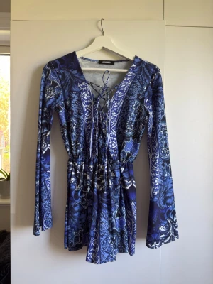 Blå mönstrad jumpsuit Missguided - Snygg blå jumpsuit från Missguided med långa ärmar och coolt paisleymönster. Djup v-ringning med snörning framtill och markerad midja. 