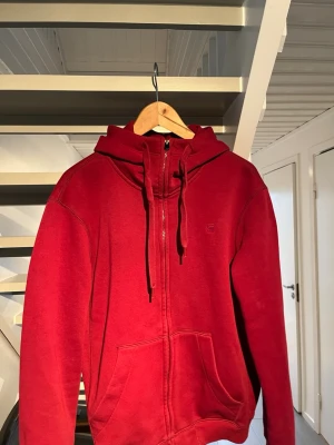 Röd zip hoodie G-Star - Snygg röd hoodie från G-Star RAW med dragkedja framtill, huva med snören och två stora fickor. Perfekt för en avslappnad streetstyle-look.  Liten logga på bröstet 