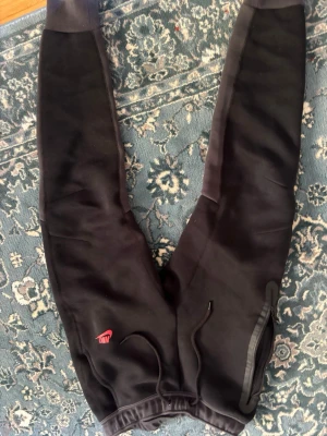 Svarta Nike mjukisbyxor med logga - Svarta mjukisbyxor från Nike med röd logga på vänster ben. Byxorna har dragsko i midjan och muddar vid bensluten. Perfekta för en avslappnad stil och tillverkade i mjukt material. Nike tech byxor 