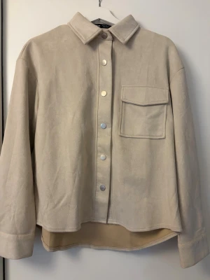 Beige kappa med ficka - Stilren beige kappa med krage och tryckknappar framtill. Kappan har en stor bröstficka med lock och är tillverkad i ett mjukt, mockaliknande material. Perfekt att bära öppen eller stängd för en avslappnad look.