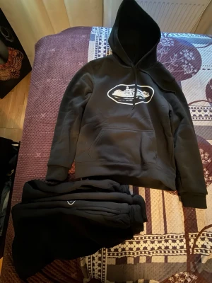 Jag säljer ett par corteiz tracksuit! - Svart hoodie med vit tryckt illustration av ett stadsmotiv på bröstet. Klassisk känguruficka framtill och justerbar huva med snören. Mjuk och skön bomullsblandning, perfekt för en avslappnad stil. Corteiz tracksuit stl S passar M