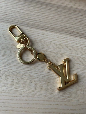 Vuitton guldfärgad nyckelring - Säljer en exklusiv guldfärgad nyckelring från  Vuitton med den ikoniska LV-logotypen. Nyckelringen har en robust karbinhake och ring, samt detaljer med ingraverad logga. Perfekt som accessoar till väska eller nycklar.
