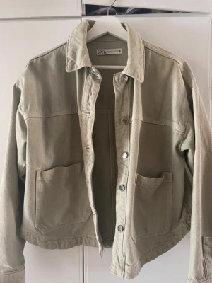 Khaki jeansjacka från ZARA - Stilren khaki färgad jeansjacka från ZARA med klassisk krage, stora framfickor och knappstängning. Jackan har en avslappnad passform och är tillverkad i kraftigt jeanstyg. Perfekt för dig som gillar en enkel och trendig look.