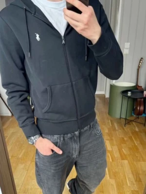 Svart zip-hoodie - Svart hoodie med dragkedja. Klassisk design med vit broderad logga på bröstet, huva med dragsko och två fickor framtill. Perfekt för en chill och stilren look.