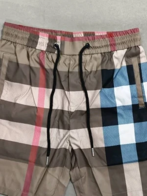 Nya Burberry shorts - Snygga burberryshorts med klassiskt rutigt mönster i beige, brunt, svart, vitt, blått och rött. Shortsen har elastisk midja med svart snörning och sidofickor. Perfekta för sommaren och ger en trendig look.