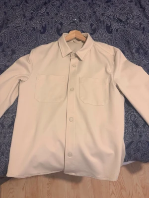 Beige overshirt från ZARA - Stilren beige overshirt från ZARA med klassisk krage, två stora bröstfickor och tryckknappar framtill. Overshirten är tillverkad i ett mjukt tyg som ger en clean och modern look. Perfekt att bära över en t-shirt för en avslappnad stil.