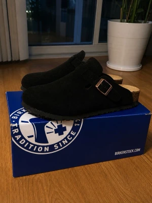 Birkenstock Boston - Svarta Birkenstock Boston tofflor i mocka med justerbart spänne i metall och klassisk korksula. Skön innersula i mocka och tidlös design som passar perfekt till både jeans och mjukisbyxor. Perfekta för dig som gillar stilrena och bekväma skor.