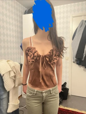 Rose satinlinnetopp  - Snygg brun linnetopp i satin med smala axelband och stora volangdetaljer framtill. Toppen har en söt knytning mellan bysten och en glansig finish som ger en lyxig känsla. Perfekt för dig som vill sticka ut med en trendig och feminin look.