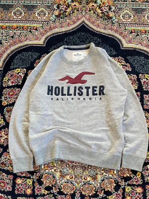 Grå sweatshirt från Hollister - Grå sweatshirt från Hollister med stor logga och texten 'California' framtill. Tröjan har rund halsringning och långa ärmar. Perfekt för en avslappnad och stilren look.