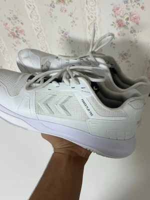 Vita Hummel Power Play Pro sneakers - Säljer ett par vita Hummel Power Play Pro sneakers med klassisk låg siluett. Skorna har meshpaneler och syntetiska detaljer för extra hållbarhet och andningsförmåga. Ikoniska Hummel-pilar på sidorna och snörning framtill. Perfekta för en sportig och clean look.