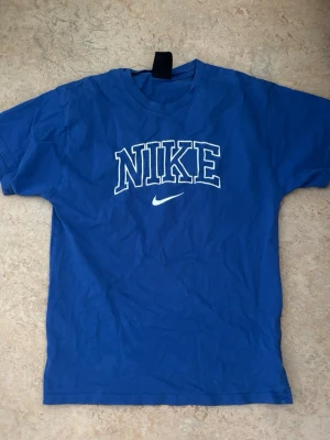 Blå Nike t-shirt med tryck - Blå t-shirt från Nike med stort vitt tryck på bröstet och klassisk logga. Modellen är kortärmad och har en avslappnad passform. Perfekt för en sportig och casual stil.