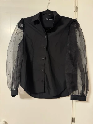 Blus/skjorta  - Stilren svart blus från Zara med klassisk krage och knäppning framtill. Ärmarna är långa och gjorda i transparent mesh med små prickar. Passar till alla tillfällen, enkel att klä upp eller ner. Endast använd en gång.