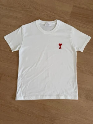 Vit t-shirt från Ami med rött hjärta - Vit t-shirt från Ami med klassisk passform och rund hals. På bröstet finns ett rött broderat hjärta med bokstaven A. Perfekt för en stilren och trendig look.