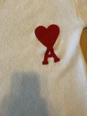 Vit stickad tröja från Ami Paris - Snygg vit stickad tröja från Ami Paris med det ikoniska röda hjärtat och bokstaven A broderat på bröstet. Tröjan har rund halsringning och ribbstickade muddar. Perfekt för dig som gillar stilrena och trendiga plagg.