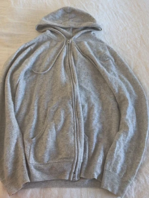 John Henric Kashmir Hoodie - En enkel grå hoodie med dragkedja och huva. Tröjan har ribbade muddar vid ärmslut och nederkant samt två fickor framtill. 