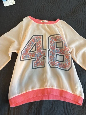 Vit och rosa topp för barn - Snygg vit topp från H&M med rosa muddar och stort blommigt 48-tryck på framsidan. Långärmad modell med transparent känsla och sportig vibe. Perfekt för dig som gillar unika detaljer och färg.
