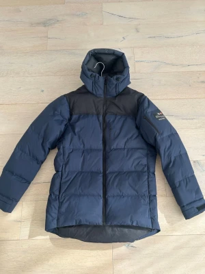 Mörkblå Sail Racing Antarctica dunjacka - Snygg mörkblå dunjacka från Sail Racing Antarctica Expedition med svarta axelpartier och huva. Jackan har dragkedja framtill, praktiska fickor och patch på ärmen. Perfekt för kalla vinterdagar när du vill hålla värmen och se cool ut. Priset går att diskutera.