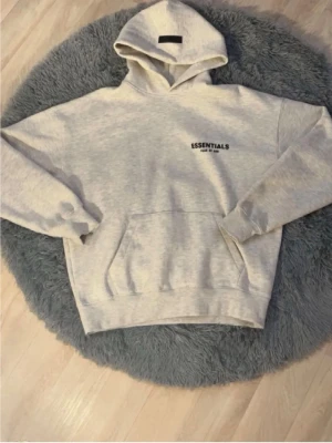 Grå Essentials Fear of God hoodie - Säljer en ljusgrå hoodie från Fear of God Essentials med svart logga på bröstet och stor text på ryggen. Hoodien har huva och magficka, samt ribbade muddar vid ärmar och nederkant. Perfekt för en avslappnad och trendig streetwear-look.