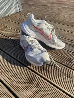 Vita Nike sneakers med röd swoosh - Snygga vita Nike sneakers med röd swoosh på sidan och vit sula. Skorna har en sportig design med meshmaterial för bra andningsförmåga och klassisk snörning. Perfekta för dig som gillar stilrena och bekväma sneakers.